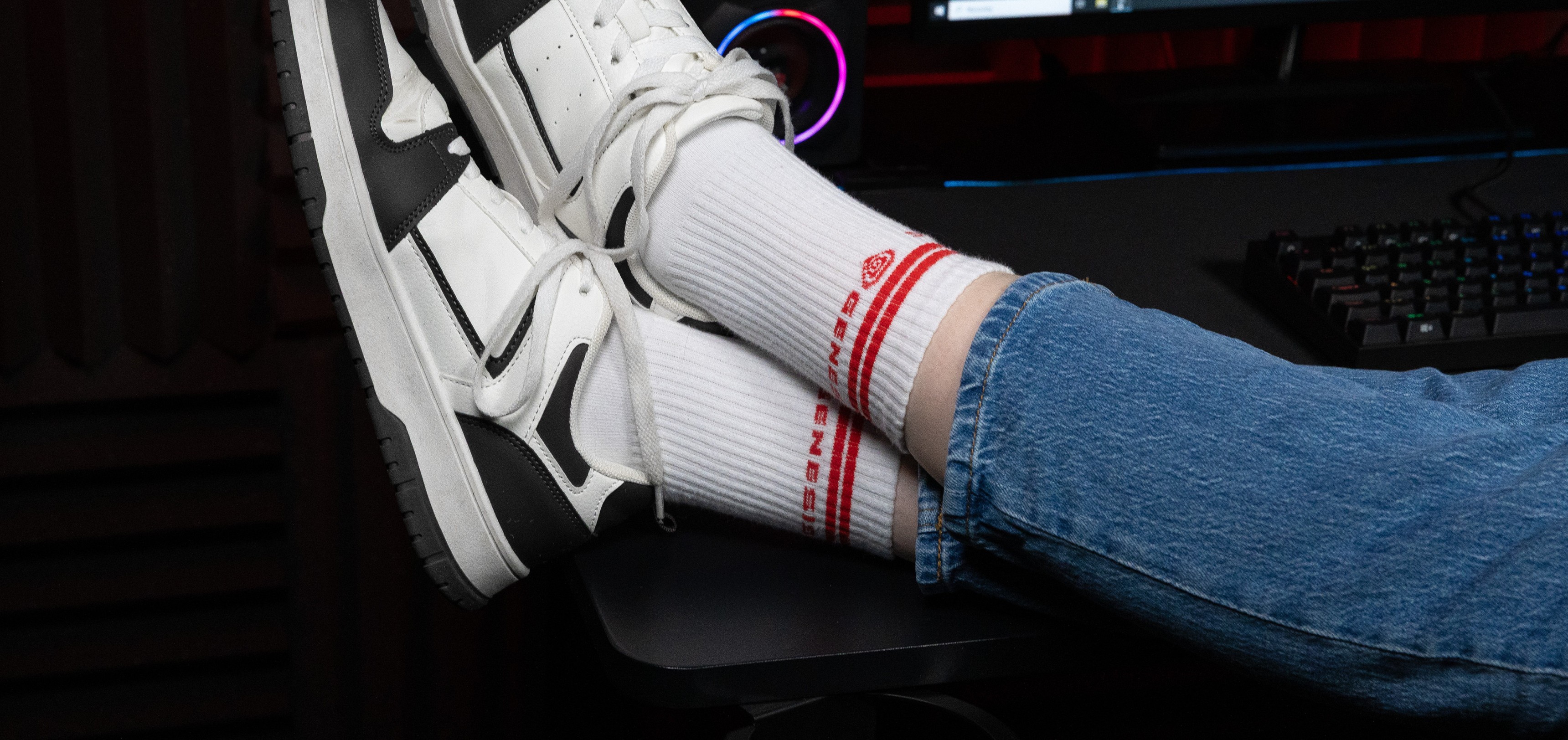 Calcetines deportivos blancos con marca roja, calzados con zapatillas blancas y negras, apoyados en un escritorio con una configuración informática