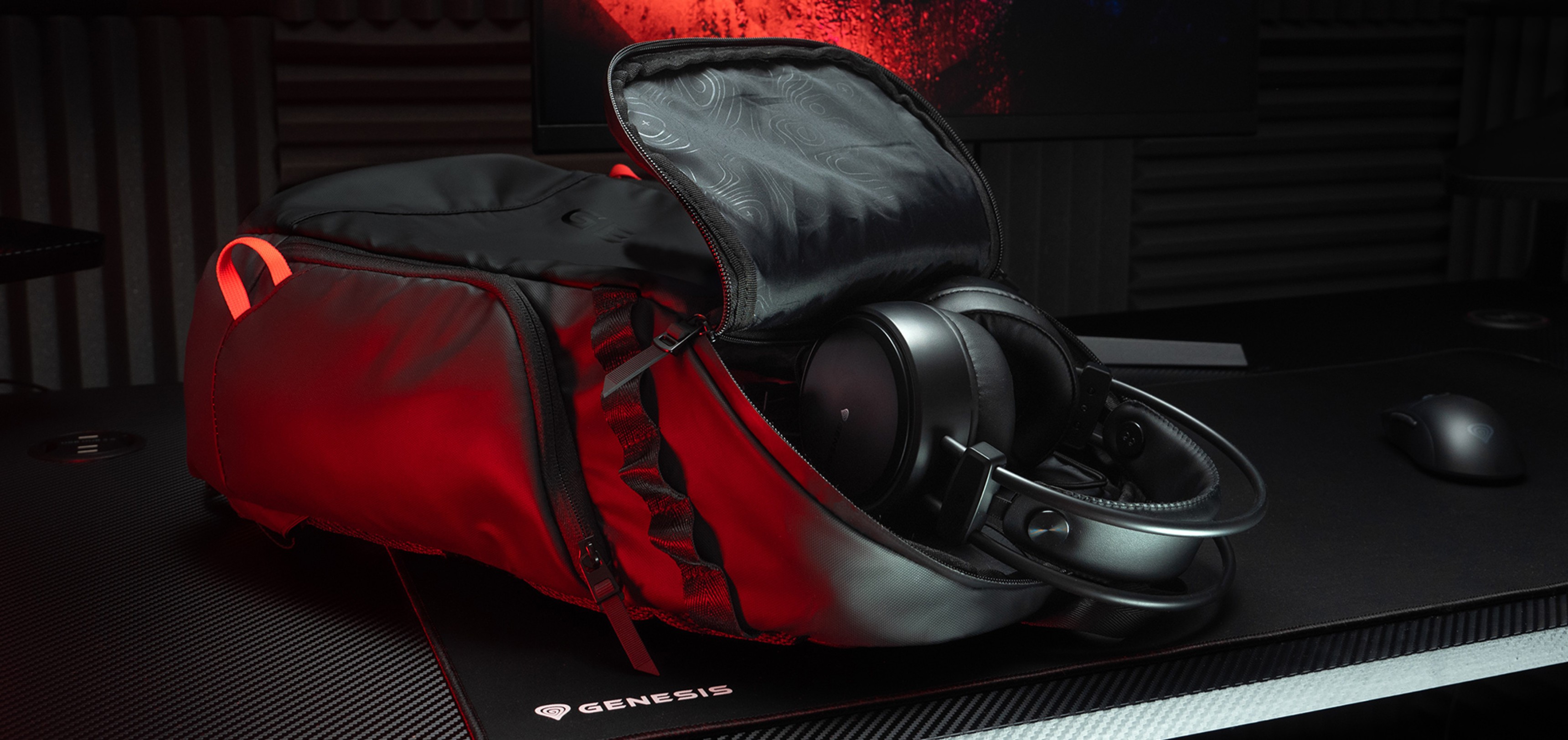 Mochila para portátil Genesis Pallad negra con un compartimento parcialmente abierto y auriculares en su interior, colocada sobre un escritorio texturizado